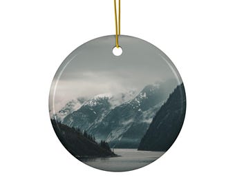 May include: A round ceramic ornament featuring a photographic print of a dark, moody mountain landscape with snow-capped peaks, misty clouds, and a calm body of water. The ornament is suspended by a thin gold string through a top hole.