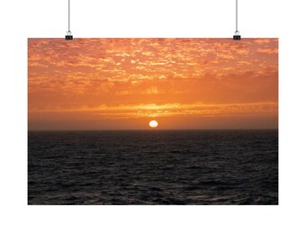 May include: A rectangular wall art print featuring a sunset over the ocean. The sun is a bright white circle on the horizon, surrounded by a vibrant orange and yellow sky filled with textured clouds above dark, rippling sea water.