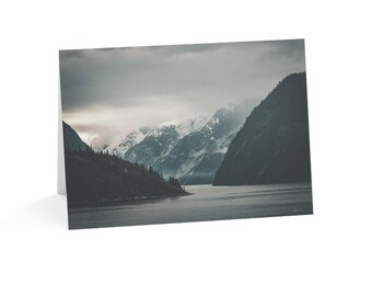 May include: A folded greeting card featuring a landscape photograph of a dark, calm body of water flanked by steep, shadowed mountains under a cloudy, overcast sky with patches of snow visible on the distant, misty mountain peaks.