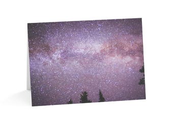 May include: A folded greeting card featuring a photograph of a night sky filled with countless stars and a purple-hued nebula. The dark silhouettes of several pine tree tops are visible along the bottom edge of the card against the starry sky.