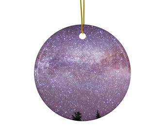 May include: A round ceramic ornament featuring a vibrant purple and pink galaxy print with a dense field of stars. The bottom edge shows the dark silhouettes of two pine trees. A thin gold string is attached to the top for hanging.