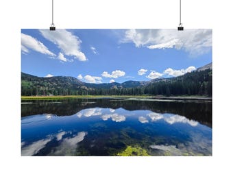 May include: A landscape wall art print featuring a calm lake reflecting a bright blue sky with scattered white clouds. The shoreline is lined with a dense forest of dark green pine trees and rolling mountains under a clear daylight sky.