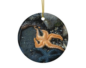 May include: A round ceramic ornament featuring a high-resolution image of a bright orange starfish resting on dark, wet rocks alongside a darker, textured starfish. The ornament is suspended by a thin gold cord attached to the top.