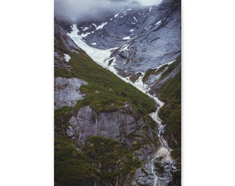 May include: A vertical landscape print featuring a rugged mountain range with dark grey rocky cliffs, patches of white snow, and deep green vegetation. A narrow, cascading stream flows down the centre of the valley under a cloudy sky.