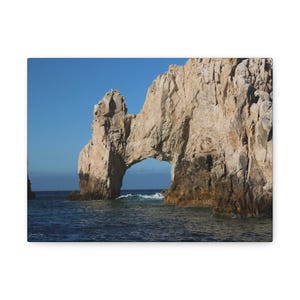 以下が含まれることがあります： 岩の多い海岸線にある、大きな開口部を持つ自然の岩のアーチの形成。アーチは、薄い茶色とベージュの岩でできており、背景には青い空と海があります。