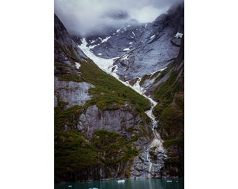 May include: A vertical wall art print featuring a dramatic mountain landscape with steep, dark grey rocky cliffs, patches of green vegetation, and a glacier flowing down the centre into a calm, dark teal body of water with small ice chunks.