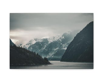 May include: A landscape wall art print featuring a dark, moody fjord scene with steep, shadowed mountains flanking a calm body of water. The peaks are partially obscured by thick, low-hanging grey clouds and patches of white snow.