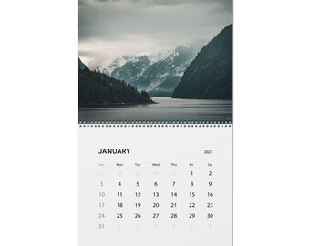 May include: A wall calendar for January 2027 featuring a top photograph of a dark, moody landscape with snow-capped mountains rising above a calm body of water under a cloudy sky. The calendar grid is printed in black on a white background.