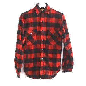 Puede incluir: Camisa de franela a cuadros rojos y negros con dos bolsillos en el pecho y cierre de botones. La camisa de manga larga está colgada en una percha blanca sobre un fondo blanco. La camisa tiene un estilo clásico de leñador.