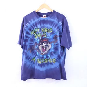 May include: A blue tie-dye t-shirt featuring the Tasmanian Devil cartoon character. The graphic includes the text "NEXT MOOD SWING 4 SECONDS" in green and white. The shirt is hanging on a wooden hanger.