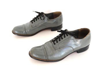 May include: A pair of gray leather oxford shoes with black laces. The shoes have a cap toe design and a dark brown sole. The shoes are displayed on a white background.