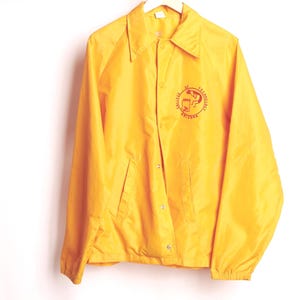 Może przedstawiać: Jasnożółta kurtka trenerska z zapięciem na guziki i kołnierzem. Kurtka ma czerwony okrągły emblemat na lewej piersi z napisem "College of Technology Arizona". Wisi na białym wieszaku.