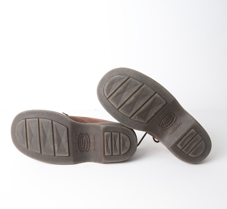 May include: A pair of brown leather lace-up shoes with a textured sole. The shoes have the word "SKECHERS" printed on the sole.