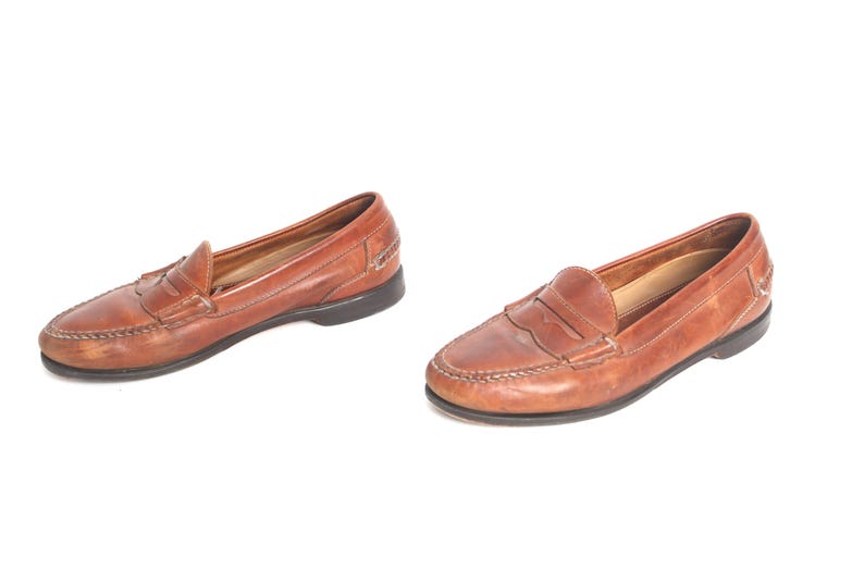 May include: A pair of brown leather loafers with a penny strap detail. The loafers have a slightly rounded toe and a low heel.