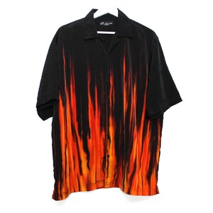 May include: Black button-down shirt with a vibrant flame design in shades of orange and red. The short-sleeved shirt is hanging on a white hanger. The shirt has a collared neckline.