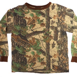 May include: Long-sleeved camouflage shirt featuring a brown collar and cuffs. The shirt's pattern includes brown tree trunks, green and brown leaves, and a tan background. This hunting-themed shirt is designed for outdoor activities.