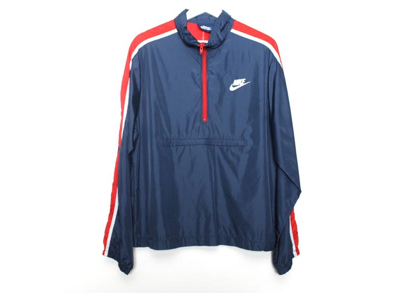 May include: Navy blue Nike windbreaker jacket with red and white stripes on the sleeves. Features a red zipper and the Nike logo in white on the chest. A classic athletic outerwear piece.