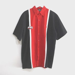 May include: A short-sleeve button-down shirt featuring a bold color-blocked design with a central red panel flanked by thin white vertical stripes and solid black side panels. The shirt includes a classic collar and a small embroidered logo patch.