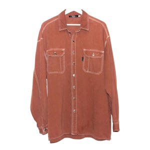 May include: A long-sleeved, button-down shirt in a reddish-brown hue. The shirt features two chest pockets and white stitching details. The collar is pointed, and the cuffs have button closures. The shirt is hanging on a white hanger.