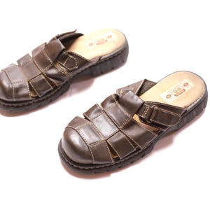 Può includere: Un paio di sandali in pelle marrone con un design intrecciato. I sandali hanno una suola spessa e scura e una soletta colorata chiara. I sandali sono lunghi circa 25 cm.