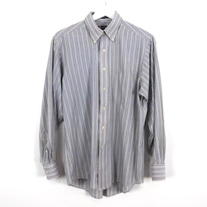 Peut inclure: Chemise à manches longues à rayures verticales bleues, grises et beiges. La chemise est suspendue à un cintre blanc. La chemise boutonnée a un col boutonné et des poignets boutonnés. Fond blanc.