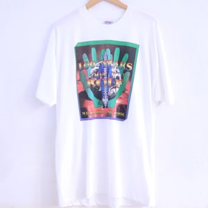 Può includere: T-shirt bianco con grafica colorata con la scritta "100 Oaks Rock & Roll" e "Manhattan, NY 1996". La grafica è inserita in un disegno a mano verde e rosso. La maglietta è appesa a una gruccia di legno.