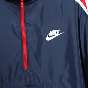 May include: Navy blue Nike windbreaker jacket with a red zipper and white logo. The jacket has a red and white stripe detail on the shoulder.