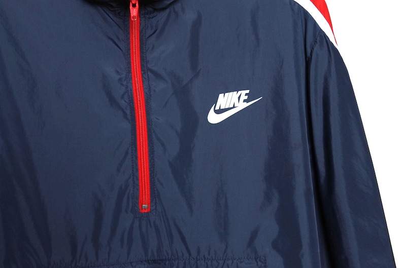 May include: Navy blue Nike windbreaker jacket with a red zipper and white logo. The jacket has a red and white stripe detail on the shoulder.