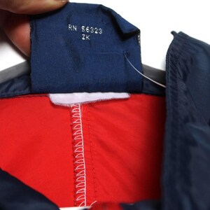 May include: Close-up of a red, white, and navy blue jacket. The jacket has a navy blue collar and a white stripe. A navy blue tag with the text "RN 56323 ZK" is visible.