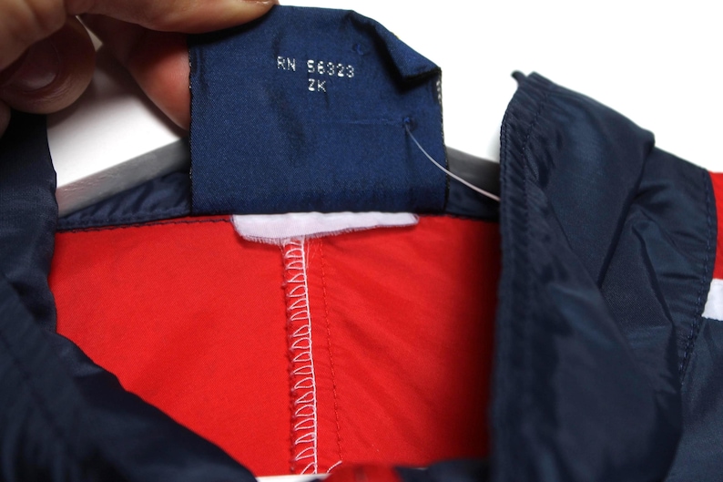 May include: Close-up of a red, white, and navy blue jacket. The jacket has a navy blue collar and a white stripe. A navy blue tag with the text "RN 56323 ZK" is visible.