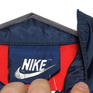 May include: Close-up of a navy blue jacket collar with a red lining. The Nike logo is prominently displayed on a navy blue tag. The jacket is hanging on a gray hanger.