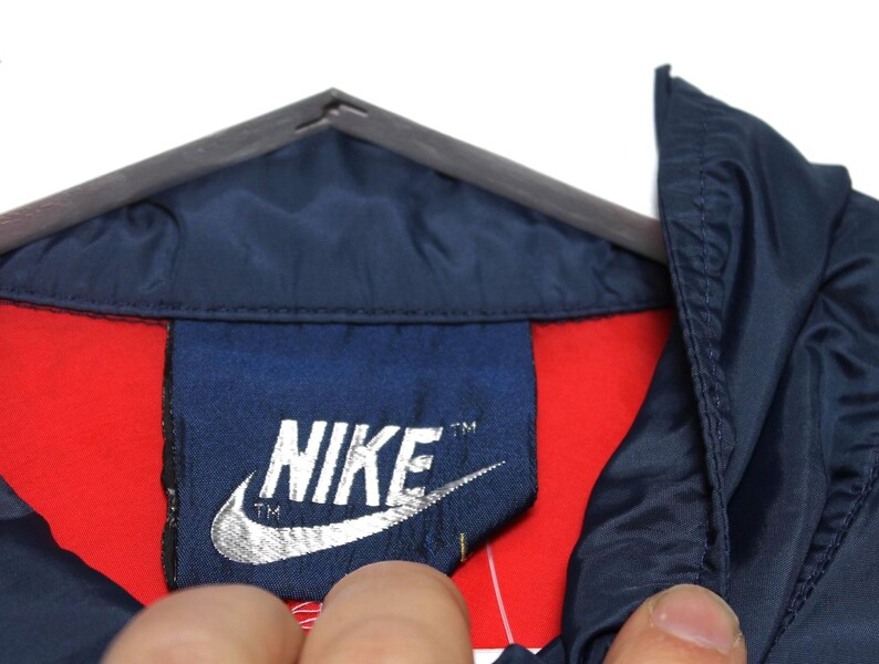 May include: Close-up of a navy blue jacket collar with a red lining. The Nike logo is prominently displayed on a navy blue tag. The jacket is hanging on a gray hanger.