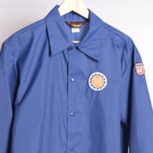 以下が含まれることがあります： ボタン留めのフロントと2つのパッチが付いた青い長袖ジャケット。1つのパッチはギアのデザインが施された円形、もう1つは「66」の数字が書かれた赤と白の盾です。