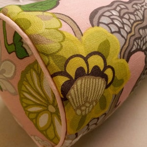 K&ouml;nnte beinhalten: Ein rosa zylindrisches Kissen mit einem Blumenmuster. Das Muster zeigt gro&szlig;e gelbe Blumen mit braunen Zentren, gr&uuml;nen Bl&auml;ttern und einem grauen und wei&szlig;en abstrakten Design.