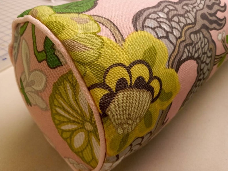 K&ouml;nnte beinhalten: Ein rosa zylindrisches Kissen mit einem Blumenmuster. Das Muster zeigt gro&szlig;e gelbe Blumen mit braunen Zentren, gr&uuml;nen Bl&auml;ttern und einem grauen und wei&szlig;en abstrakten Design.