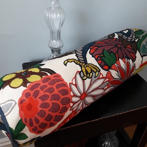 K&ouml;nnte beinhalten: Ein zylindrisches Kissen mit wei&szlig;em Hintergrund und einem bunten Blumenmuster. Das Muster zeigt gro&szlig;e rote, gelbe und gr&uuml;ne Blumen mit schwarzen Umrissen. Das Kissen liegt auf einem schwarzen Tisch.