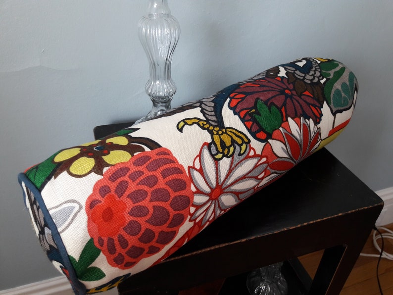 K&ouml;nnte beinhalten: Ein zylindrisches Kissen mit wei&szlig;em Hintergrund und einem bunten Blumenmuster. Das Muster zeigt gro&szlig;e rote, gelbe und gr&uuml;ne Blumen mit schwarzen Umrissen. Das Kissen liegt auf einem schwarzen Tisch.