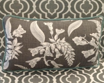 May include: Rectangular decorative pillow with a dark gray background and a white floral pattern. The pillow has a teal green border and is placed on a chair with a gray and white geometric design. The pillow's design features stylized flowers and leaves.