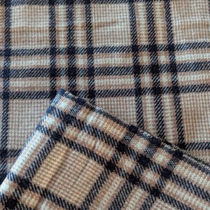 Puede incluir: Primer plano de una tela a cuadros beige y negra. La tela presenta un patrón clásico de líneas que se cruzan, creando un diseño de cuadrícula. La tela está doblada, revelando el borde del material.