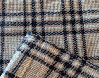 May include: Close-up of a tan and black plaid fabric. The fabric has a classic pattern of intersecting lines, creating a grid-like design. The fabric is folded over, revealing the edge of the material.