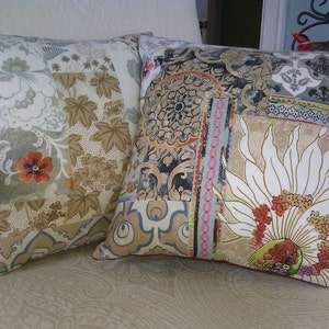 Pode incluir: Duas almofadas decorativas com designs de patchwork. Uma almofada apresenta padrões florais e de folhas em bege, cinza e laranja. A outra tem uma grande flor branca e vários padrões geométricos em cores semelhantes.