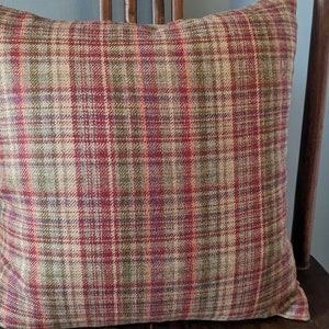 May include: Square decorative pillow with a plaid pattern in shades of red, green, and beige. The pillow is placed on a wooden chair, showcasing its textured fabric and color palette. A cozy home decor item.