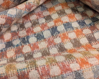 May include: Close-up of a woven fabric with a checkered pattern. The fabric features squares in shades of orange, blue, brown, and cream. The material is folded, showing the texture and weave of the fabric.