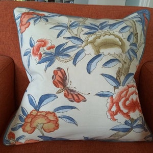 Op de afbeelding: Een vierkant sierkussen met een gebroken witte achtergrond en een printpatroon van oranje en beige pioenrozen, blauwe bladeren en een centrale oranje vlinder. Het kussen is afgewerkt met een blauwe bies langs de rand van de stof.