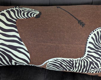 May include: Brown rectangular decorative pillow featuring two zebras in black and white stripes. One zebra is upright, and the other is lying down. A black arrow is also on the pillow.