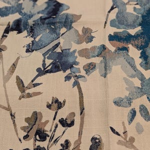Puede incluir: Primer plano de una tela con un estampado floral. El diseño presenta flores y hojas de estilo acuarela azules, turquesas y marrones sobre un fondo color crema. La tela tiene un aspecto texturizado.