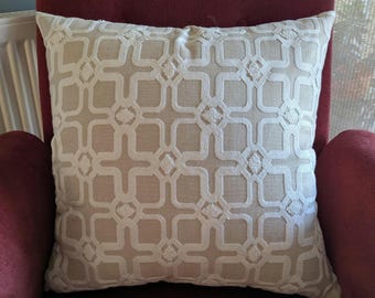 May include: A square beige pillow with a white geometric pattern. The pillow has a textured surface and is placed on a red chair. The pattern consists of interconnected squares and diamond shapes, creating a decorative design.