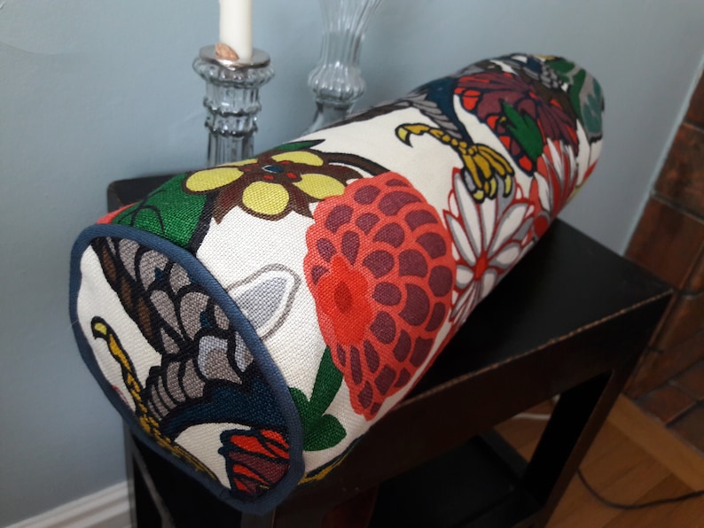 K&ouml;nnte beinhalten: Ein zylindrisches Kissen mit wei&szlig;em Hintergrund und einem bunten Blumenmuster. Das Muster zeigt rote, orange, gelbe, gr&uuml;ne, blaue und schwarze Blumen und Bl&auml;tter. Das Kissen hat einen dunkelblauen Saum an den R&auml;ndern.