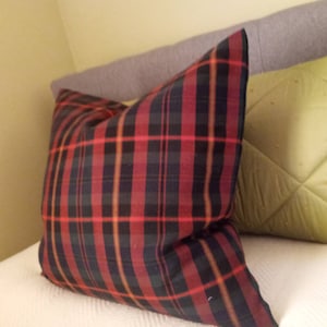 May include: A square throw pillow featuring a classic plaid pattern with intersecting lines in shades of deep red, black, and dark green, accented by thin, bright orange stripes that create a bold, structured grid across the entire fabric surface.