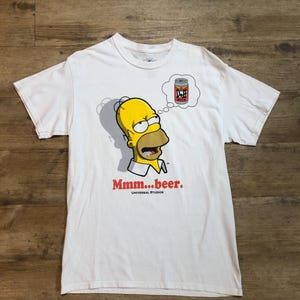 Könnte beinhalten: Weißes T-Shirt mit einem Cartoon-Bild von Homer Simpson, der Figur aus der Simpsons-Fernsehserie, mit einer Gedankenblase über seinem Kopf, die eine Dose Duff Beer zeigt. Der Text "Mmm...beer." ist unter dem Bild gedruckt. Der Text "Universal Studios" ist unter dem Text "Mmm...beer." gedruckt.